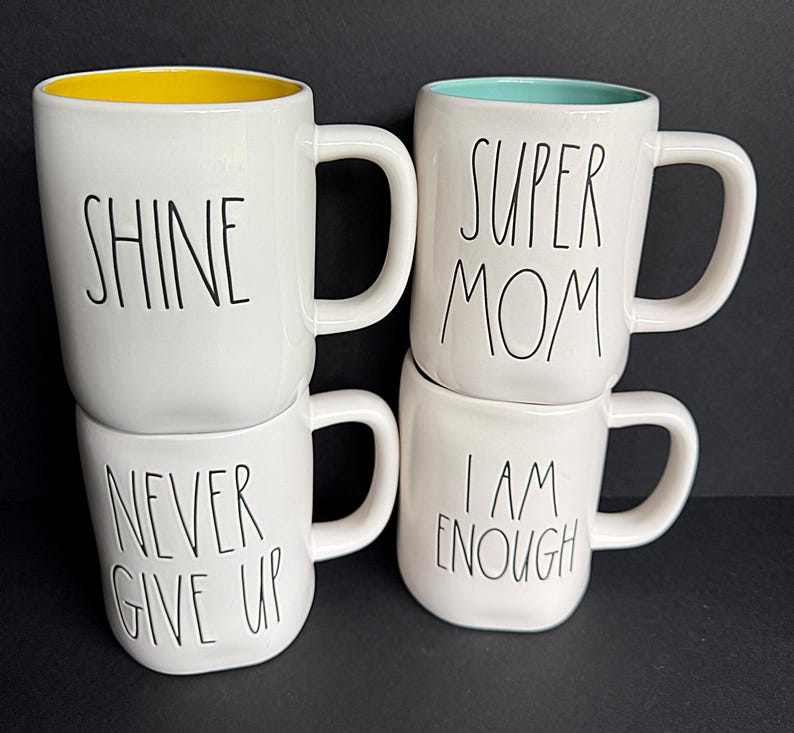 May include: Four white ceramic mugs with black lettering. The top left mug has a yellow interior and says SHINE. The bottom left mug says NEVER GIVE UP. The top right mug has a teal interior and says SUPER MOM. The bottom right mug says I AM ENOUGH.