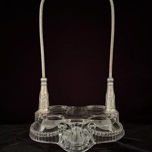 May include: An antique clear glass serving piece with a silver-toned handle. The glass base has a decorative beaded edge and a central, ornate design. The handle arches over the top, connecting to the base with detailed silver supports.