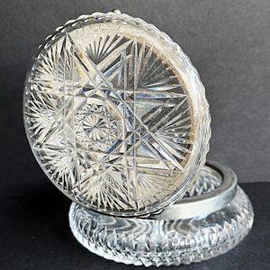 May include: A clear, cut-glass trinket box with a starburst pattern on the lid. The lid is open, revealing the interior of the box. The box has a silver-colored metal rim and base. The glass is highly reflective.