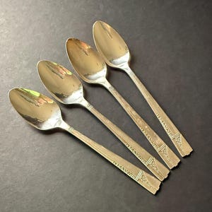May include: Four vintage silver spoons arranged diagonally. The spoons have oval bowls and ornate, rectangular handles with a decorative pattern. The spoons are made of a shiny, reflective metal, likely silver or a silver-colored alloy.