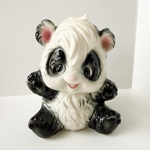 May include: A ceramic panda figurine with a glossy finish. The panda has black and white fur, black eyes, and pink accents on its ears and mouth. It is sitting with its arms outstretched, giving it a friendly appearance.
