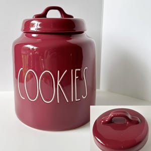 May include: A deep burgundy ceramic cookie jar with a matching lid featuring a handle. The word COOKIES is embossed in white, stylized, thin, uppercase lettering on the front of the jar. A separate view of the lid is shown in the bottom corner.