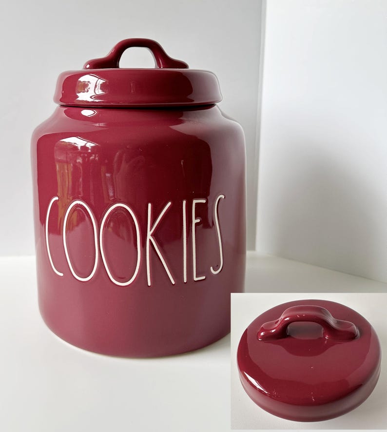 May include: A deep burgundy ceramic cookie jar with a matching lid featuring a handle. The word COOKIES is embossed in white, stylized, thin, uppercase lettering on the front of the jar. A separate view of the lid is shown in the bottom corner.