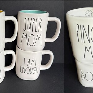 May include: A collection of five white ceramic mugs with black handwritten-style text. The mugs feature the phrases SHINE, NEVER GIVE UP, SUPER MOM, I AM ENOUGH, PINCH ME, and BOO. One mug has a yellow interior, one has a teal interior, and one has a clover icon.