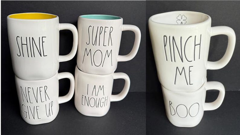 May include: A collection of five white ceramic mugs with black handwritten-style text. The mugs feature the phrases SHINE, NEVER GIVE UP, SUPER MOM, I AM ENOUGH, PINCH ME, and BOO. One mug has a yellow interior, one has a teal interior, and one has a clover icon.