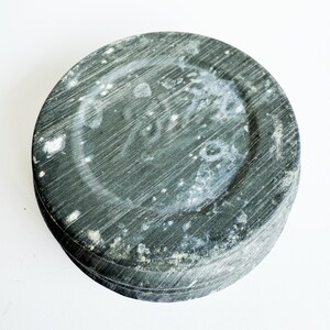May include: A round, dark gray stone object with a textured surface and a circular indentation in the center. The stone has visible striations and white patches, suggesting age or wear. The object is set against a white background.