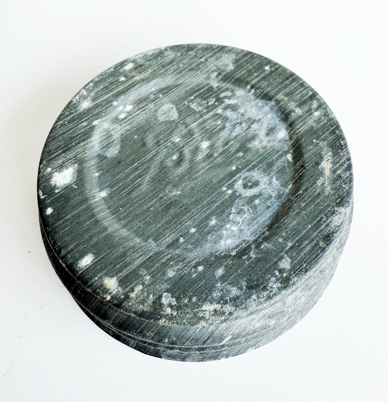 May include: A round, dark gray stone object with a textured surface and a circular indentation in the center. The stone has visible striations and white patches, suggesting age or wear. The object is set against a white background.