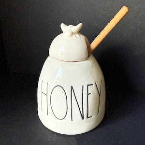 May include: A white ceramic honey pot with a rounded lid featuring a small sculpted bee on top. The word HONEY is printed in black, tall, thin, sans-serif capital letters on the front. A light brown wooden honey dipper rests inside the pot.