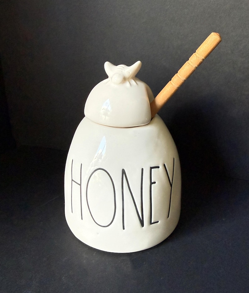 May include: A white ceramic honey pot with a rounded lid featuring a small sculpted bee on top. The word HONEY is printed in black, tall, thin, sans-serif capital letters on the front. A light brown wooden honey dipper rests inside the pot.