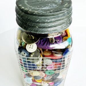 May include: A clear glass jar filled with a variety of colorful buttons. The jar has a textured, dark gray lid and a decorative, grid-like pattern on the lower half. Buttons are in various sizes, shapes, and colors.