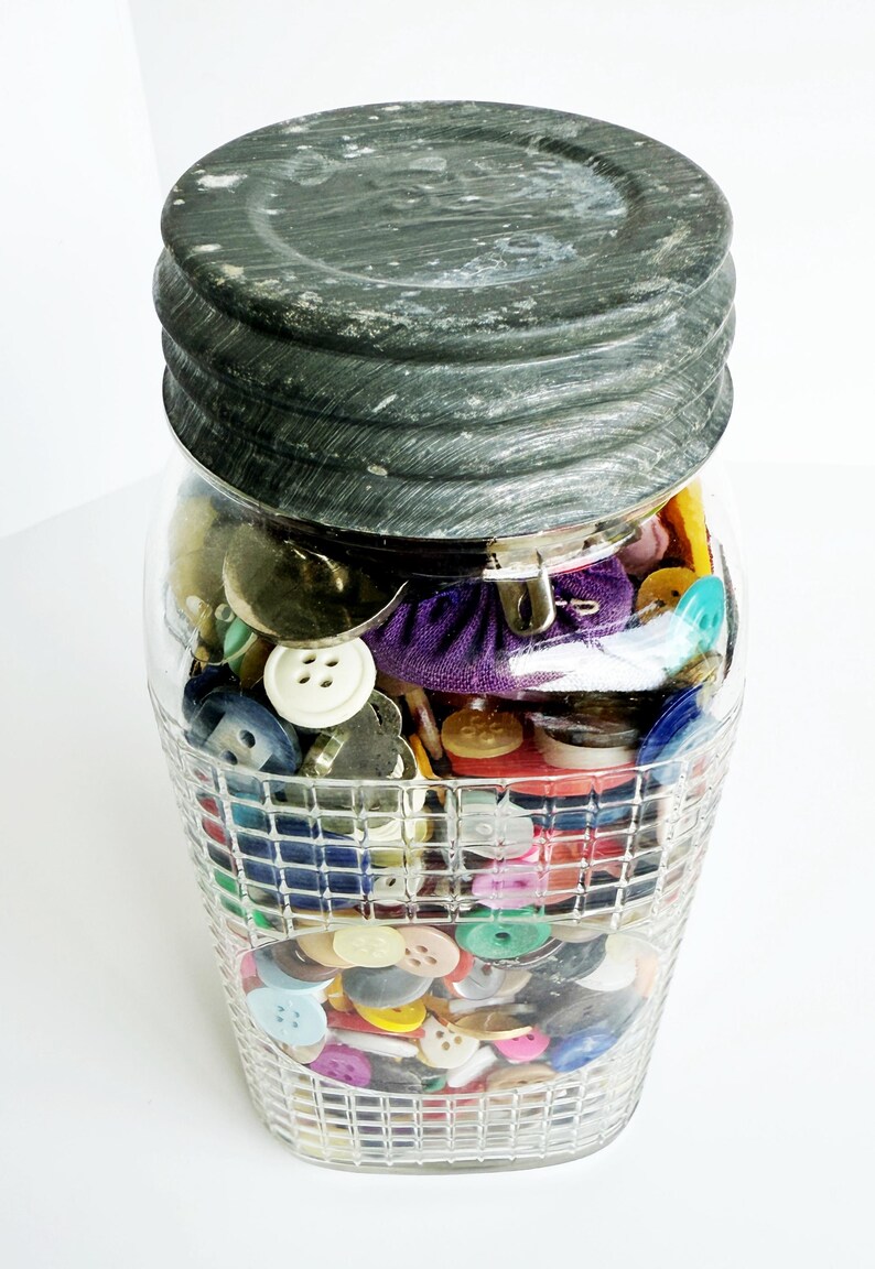 May include: A clear glass jar filled with a variety of colorful buttons. The jar has a textured, dark gray lid and a decorative, grid-like pattern on the lower half. Buttons are in various sizes, shapes, and colors.