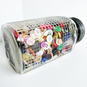May include: A clear glass jar filled with a variety of colorful buttons. The jar has a textured, grid-like pattern and a black lid. The buttons are in various colors, including red, pink, yellow, green, and blue, and are of different sizes and shapes.