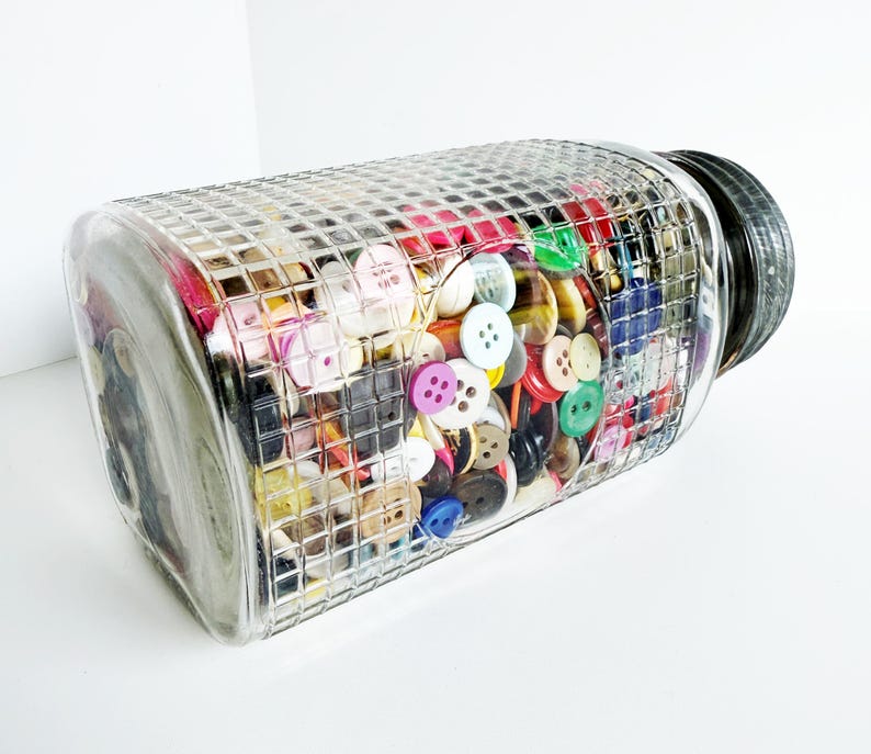 May include: A clear glass jar filled with a variety of colorful buttons. The jar has a textured, grid-like pattern and a black lid. The buttons are in various colors, including red, pink, yellow, green, and blue, and are of different sizes and shapes.
