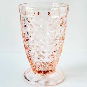 May include: A vintage pink glass with a diamond pattern. The glass has a pedestal base and a flared rim. The pink color is soft and translucent, and the glass appears to be in good condition. This is a decorative glassware item.