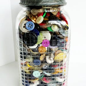 May include: A clear glass jar filled with a variety of colorful buttons. The jar has a textured, dark gray lid and a grid-like pattern on the glass. The buttons are in various sizes, shapes, and colors, including blue, yellow, green, and white.