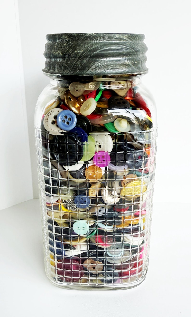May include: A clear glass jar filled with a variety of colorful buttons. The jar has a textured, dark gray lid and a grid-like pattern on the glass. The buttons are in various sizes, shapes, and colors, including blue, yellow, green, and white.