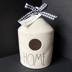 May include: A cream-colored ceramic birdhouse with a cylindrical body and a conical top. The word HOME is printed in black lettering on the front below a circular opening. A black and white gingham ribbon is tied at the top with a Rae Dunn tag.