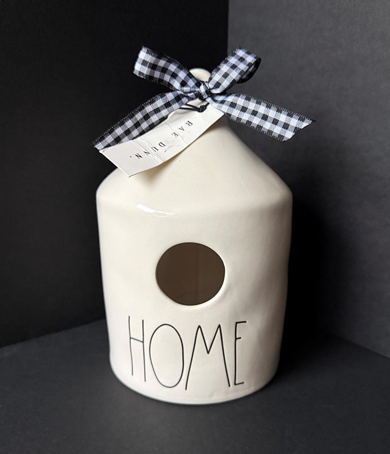May include: A cream-colored ceramic birdhouse with a cylindrical body and a conical top. The word HOME is printed in black lettering on the front below a circular opening. A black and white gingham ribbon is tied at the top with a Rae Dunn tag.
