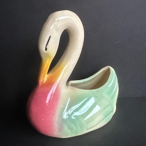 May include: A ceramic swan-shaped planter with a curved neck and a small opening for plants. The swan is painted with a gradient of colors, including pink, green, yellow, and white. The planter is designed to sit upright.