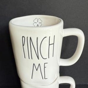 May include: A white ceramic mug with a glossy finish featuring the black text PINCH ME written in a tall, thin, handwritten font. Inside the upper rim of the mug, there is a small, simple black outline of a four-leaf clover icon.