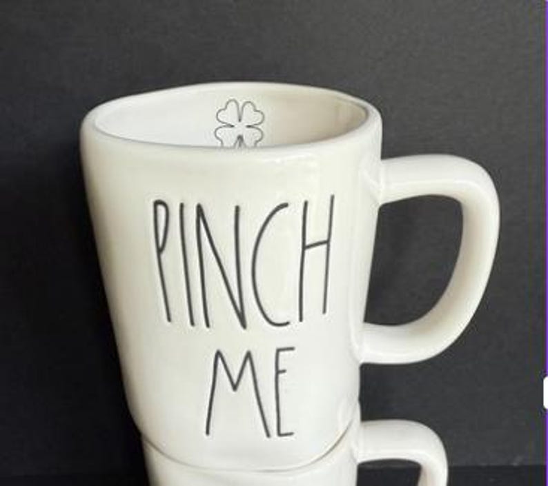 May include: A white ceramic mug with a glossy finish featuring the black text PINCH ME written in a tall, thin, handwritten font. Inside the upper rim of the mug, there is a small, simple black outline of a four-leaf clover icon.