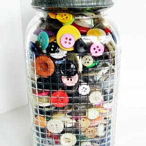 May include: A clear glass jar filled with a variety of colorful buttons. The buttons are in various sizes, shapes, and colors, including yellow, pink, blue, and black. The jar has a textured, grid-like pattern and a dark gray lid.