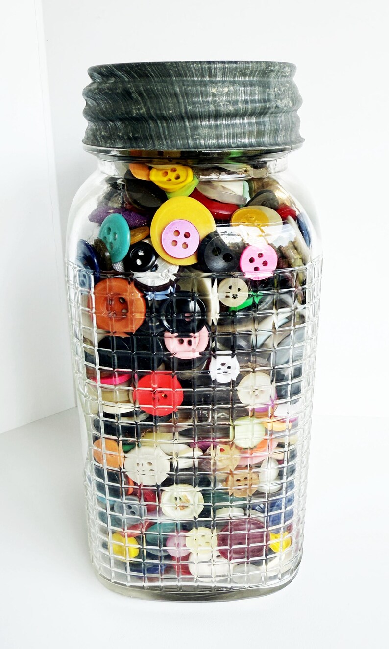 May include: A clear glass jar filled with a variety of colorful buttons. The buttons are in various sizes, shapes, and colors, including yellow, pink, blue, and black. The jar has a textured, grid-like pattern and a dark gray lid.
