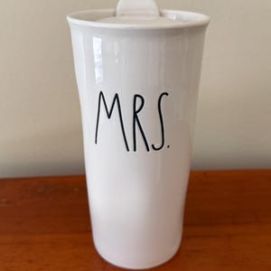 May include: A white ceramic travel mug with a lid, featuring the word MRS. printed in black, handwritten-style lettering on the front. The mug has a slightly tapered cylindrical shape with a smooth, glossy finish and a simple, clean design.
