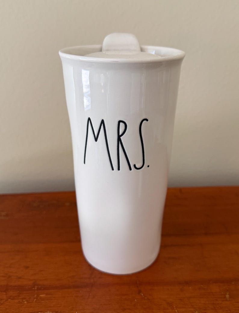 May include: A white ceramic travel mug with a lid, featuring the word MRS. printed in black, handwritten-style lettering on the front. The mug has a slightly tapered cylindrical shape with a smooth, glossy finish and a simple, clean design.