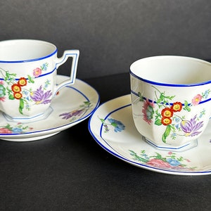 May include: Two white teacups and saucers with a floral design. The cups and saucers have a blue trim and a geometric shape. The floral design includes red, purple, and yellow flowers.