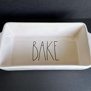 May include: A rectangular off-white ceramic baking dish with integrated side handles. The interior base features the word BAKE written in a tall, thin, black sans-serif font. The dish has a smooth, glossy finish and a deep, open basin design.