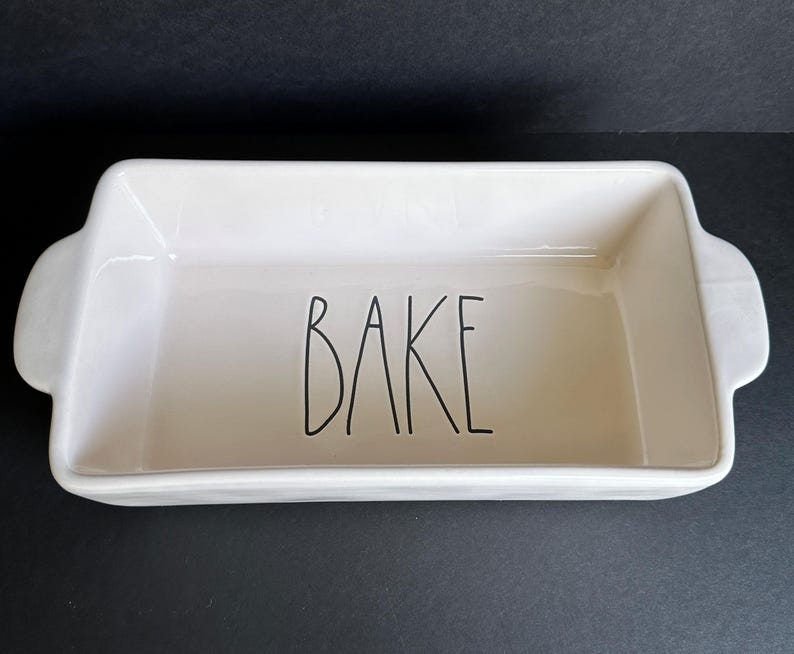 May include: A rectangular off-white ceramic baking dish with integrated side handles. The interior base features the word BAKE written in a tall, thin, black sans-serif font. The dish has a smooth, glossy finish and a deep, open basin design.