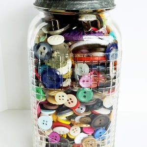May include: A clear glass jar filled with a variety of colorful buttons. The buttons are in various sizes, shapes, and colors, including blue, green, red, and yellow. The jar has a dark gray metal lid.