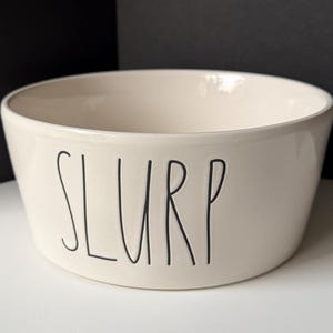 May include: A cream-colored ceramic bowl with a smooth, glossy finish. The word SLURP is printed on the front in a tall, thin, black sans-serif font. The bowl has a wide, open top and a slightly tapered base, presenting a clean, simple design.