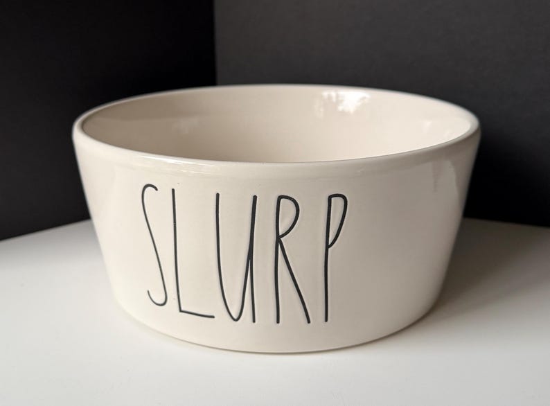 May include: A cream-colored ceramic bowl with a smooth, glossy finish. The word SLURP is printed on the front in a tall, thin, black sans-serif font. The bowl has a wide, open top and a slightly tapered base, presenting a clean, simple design.