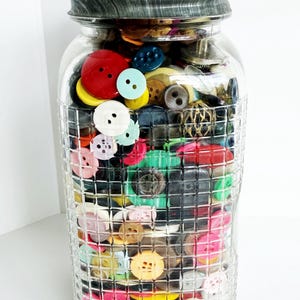 May include: A clear glass jar filled with a variety of colorful buttons. The buttons are in various sizes and colors, including red, yellow, blue, and pink. The jar has a textured, grid-like pattern and a gray metal lid.