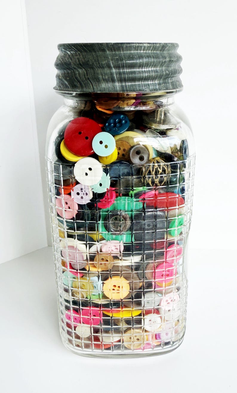 May include: A clear glass jar filled with a variety of colorful buttons. The buttons are in various sizes and colors, including red, yellow, blue, and pink. The jar has a textured, grid-like pattern and a gray metal lid.