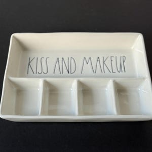 May include: A rectangular off-white ceramic tray with a glossy finish, featuring four distinct compartments. The upper section is a wide, open space with the black text KISS AND MAKEUP printed in a simple, hand-lettered style font.
