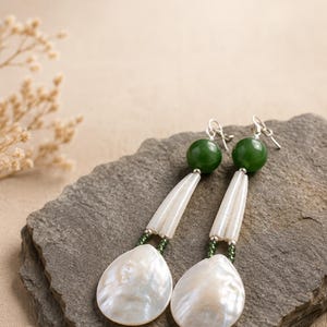 May include: A pair of drop earrings featuring a round dark green bead at the top, followed by two slender white cylindrical pieces, and finished with a large, flat, pear-shaped white shell pendant. The components are connected by small green beads.