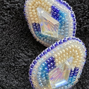 May include: Handmade beaded earrings with a geometric design. The earrings feature a central iridescent crystal, surrounded by cream, light blue, and dark blue beads. The edges are outlined with dark blue beads. A white bead hangs below.