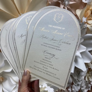 May include: A stack of ivory wedding ceremony programs with silver foil accents. The programs are shaped like arches and feature the text "The Wedding of Mariza Mwana & Robert James Calahan" and the date "Saturday, December 19, 2020".
