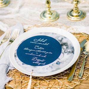 May include: A white plate with a blue and gray watercolor design featuring a crescent moon. The plate has a blue circle with white text that reads "Salad, serreno engar salad, Entrie, filet migasm with potato gratin, mrendamin make make, Dessert, wedding enke".