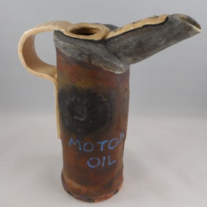 May include: A ceramic pitcher shaped like an oil can with a brown, rusty exterior. The pitcher has a handle and a spout. The words "MOTOR OIL" are painted in blue on the front of the pitcher.