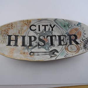 May include: White ceramic oval dish with a brown, white, and green design. The dish has the words "City Hipster" written in black.
