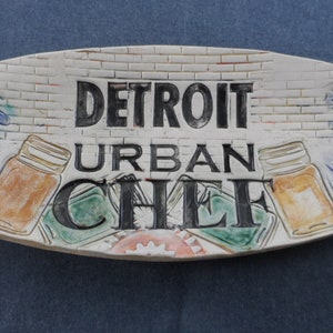 May include: White ceramic oval serving dish with a brick pattern and the words "Detroit Urban Chef" in black lettering. The dish is decorated with blue and green floral designs and two jars with orange lids.