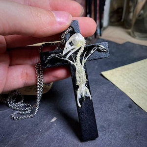 May include: A black cross pendant featuring a bird skeleton design, suspended from a silver chain. The bird's skull and bones are visible against the black cross, creating a gothic aesthetic.