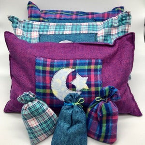 May include: Three purple, blue, and green plaid pillow covers with a white crescent moon and star applique. The pillows are arranged in a stack with the purple pillow on top. There are three small plaid bags in front of the pillows.