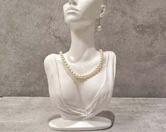 May include: A white ceramic bust of a woman with a pearl necklace and a single pearl earring. The bust is a decorative vase with a hollowed-out head. The bust is on a grey surface.