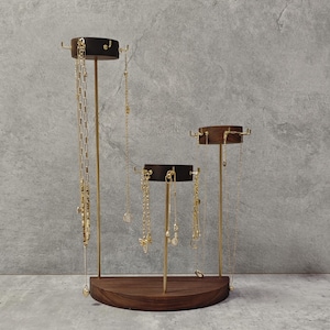 Puede incluir: Un expositor de joyas de tres niveles con base y plataformas de madera oscura. El expositor presenta soportes metálicos de color latón y ganchos para colgar collares. Se exhiben varios collares dorados en el expositor.