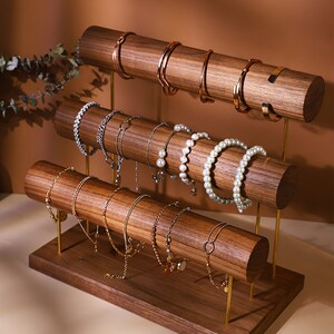 May include: A three-tiered jewellery display stand made of dark wood with gold-coloured metal supports. The stand holds various bracelets, including gold bangles, beaded bracelets, and pearl strands. The wooden rods are cylindrical.